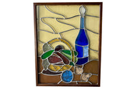 Framed stained glass still-life of a basket of vegetable and wine bottle.