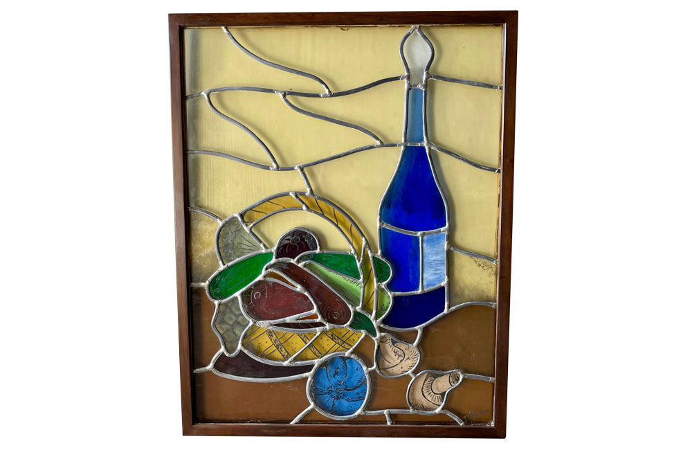 Framed stained glass still-life of a basket of vegetable and wine bottle.