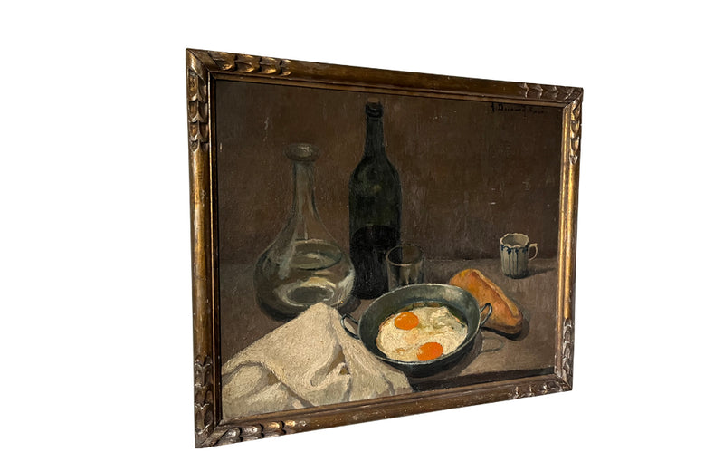 Lovely, French still life painting of fried eggs in a pan, a carafe of water, bottle of wine, glass, loaf of bread, ceramic cup and cloth napkin. 